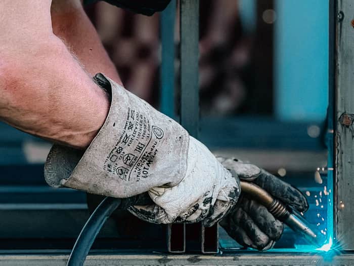 Hand im Schweißerhandschuh setzt mit dem Brenner eine Naht an einem Stahlrahmen, Funken sprühen