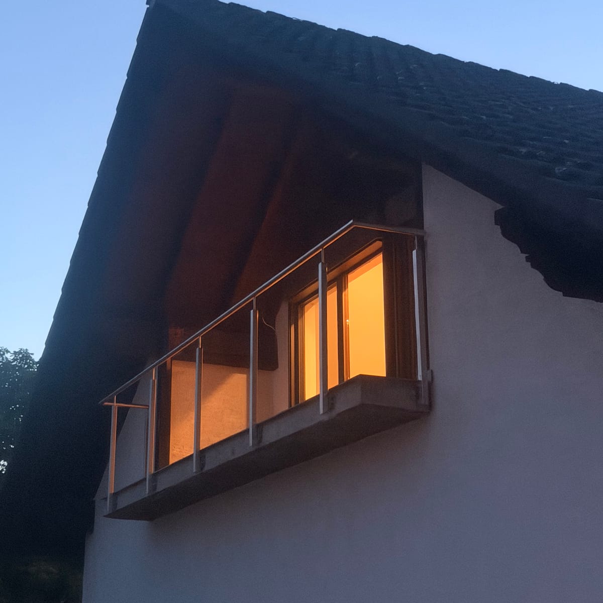 Balkon unter dem Dachüberstand eines Wohnhauses in der Dämmerung, das Geländer besteht aus Edelstahlpfosten mit Glasfüllung