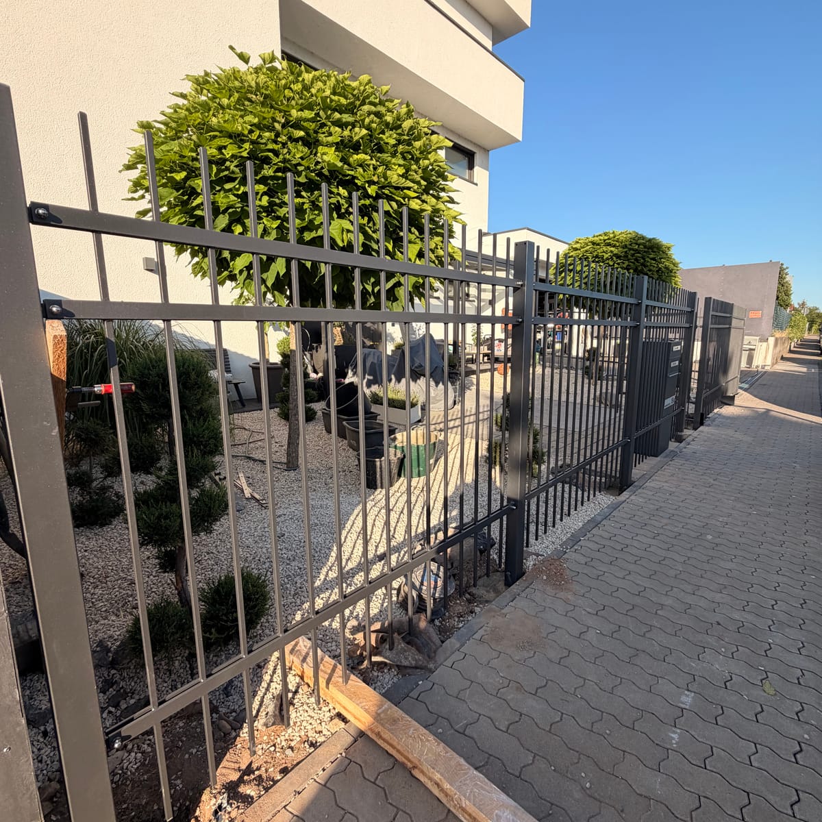 Langer anthrazitfarbener Stabzaun entlang einer gepflasterten Einfahrt vor einem modernen Wohnhaus, im Vordergrund liegt noch Montagematerial